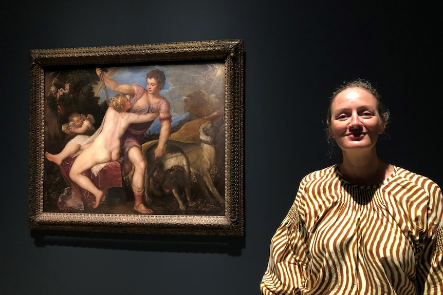 A smiling woman stands in front of a romantic painting by Titian