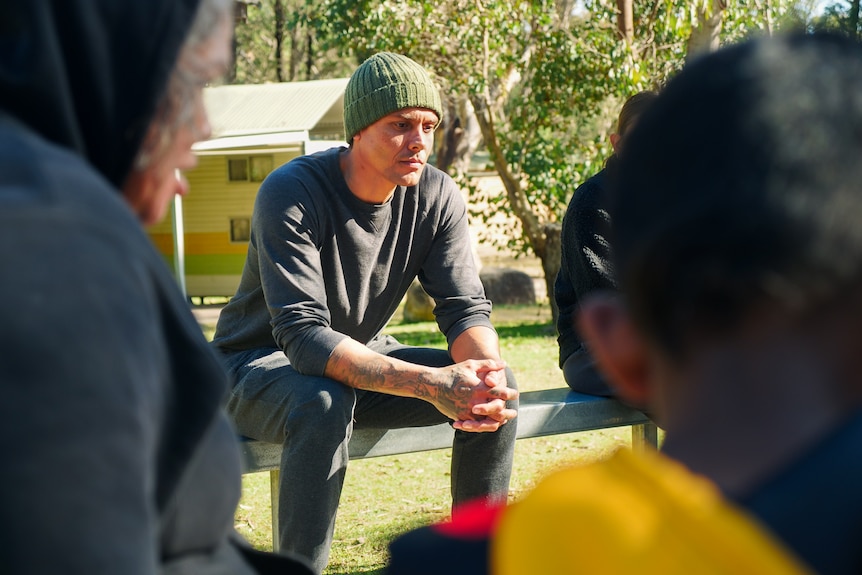 A young Indigenous man in a beanie sits on a bench looking contemplative.