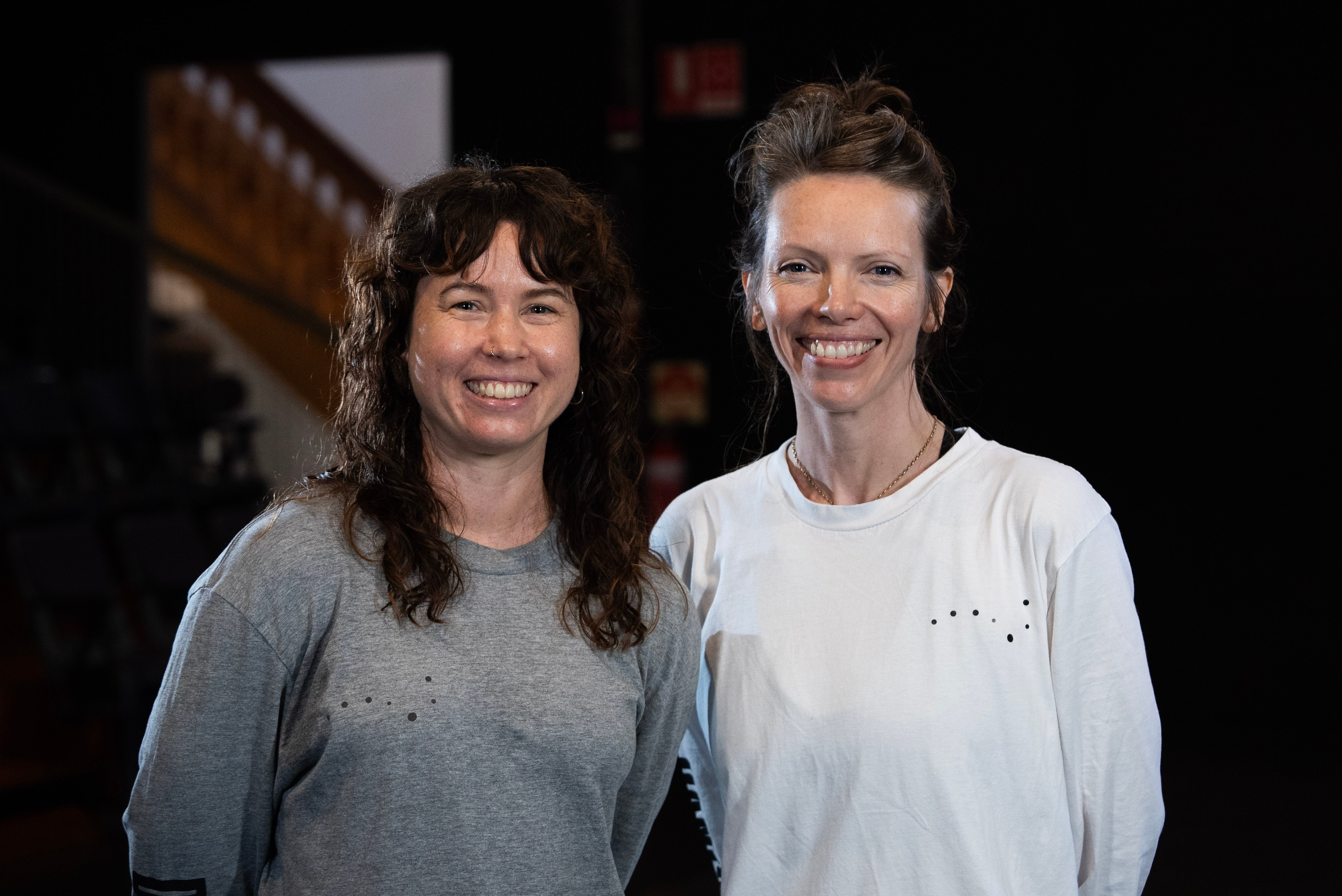 A woman in a grey top with shoulder legnth curly hair and a woman in a white shirt with her hair up - both smiling. 