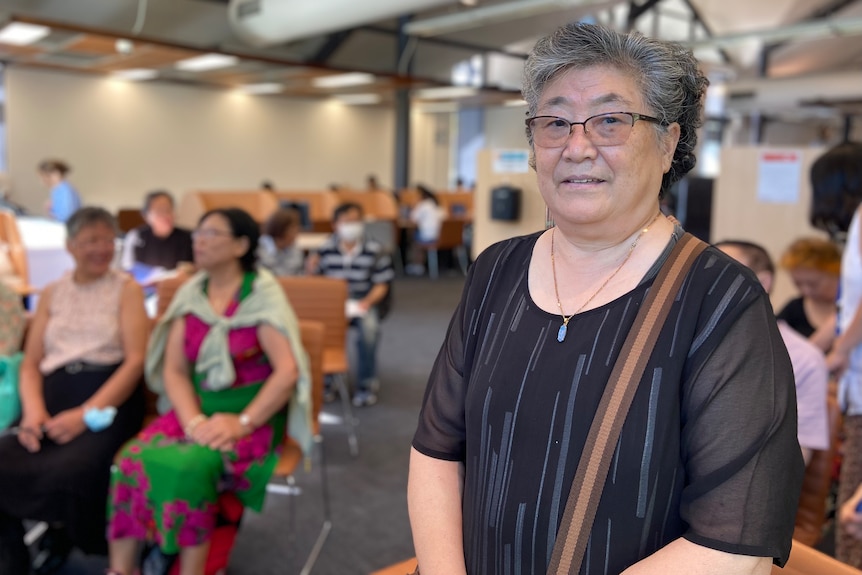 An elderly woman in a black top and glasses standing in a hall smiling at the camera