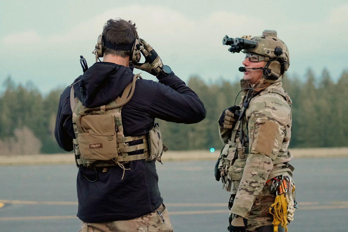 Two men in military combat gear on a tarmac: one has his back to the camera, the other is side-on in a helmet.