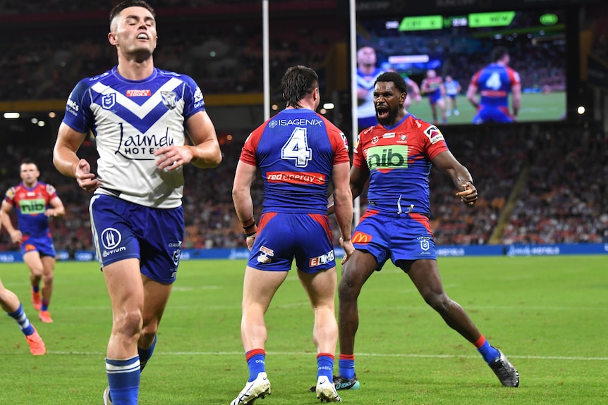 A Bulldogs player closes his eyes in dejection as two Newcastle players celebrate a try in the background.