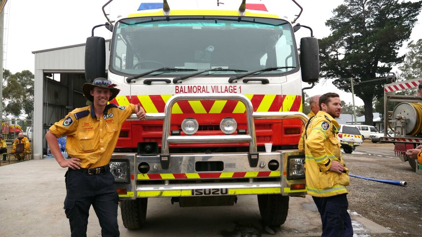 The front of a firetruck parked at a station, with two men in uniform leaning on either side of it.