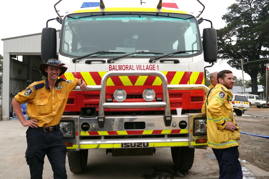 The front of a firetruck parked at a station, with two men in uniform leaning on either side of it.