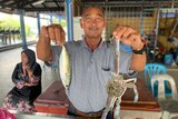 A man in a Malaysian fish market holds up a fish and a crab