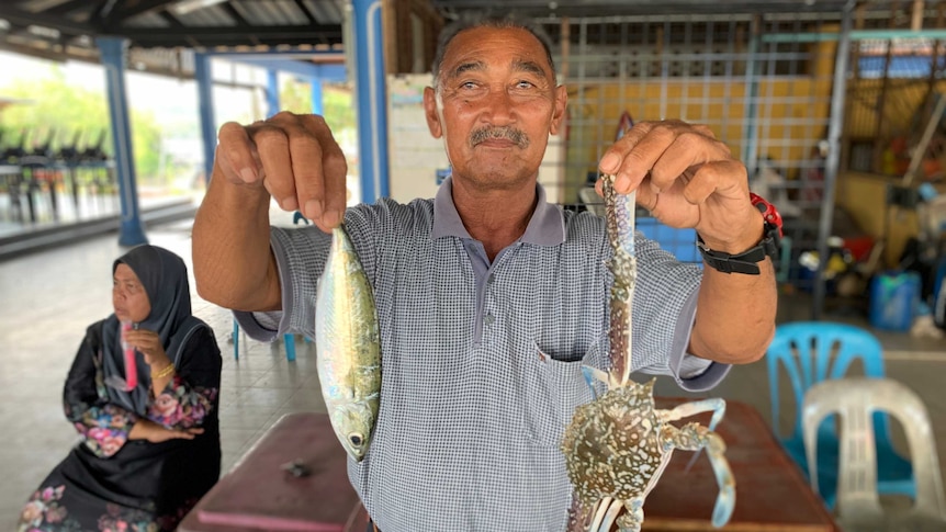 A man in a Malaysian fish market holds up a fish and a crab