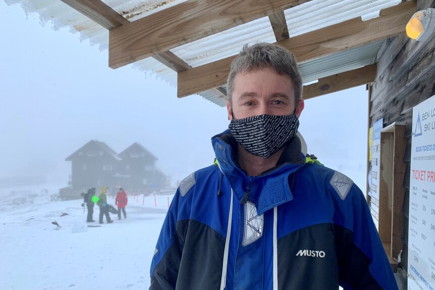 A man wearing a face mask stands under a veranda with a group of skiers in the snow in the background