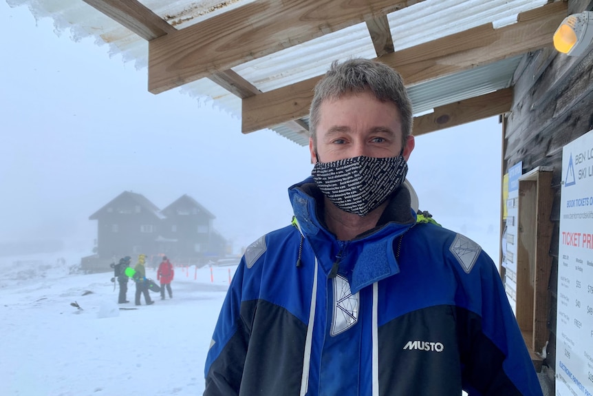 A man wearing a face mask stands under a veranda with a group of skiers in the snow in the background