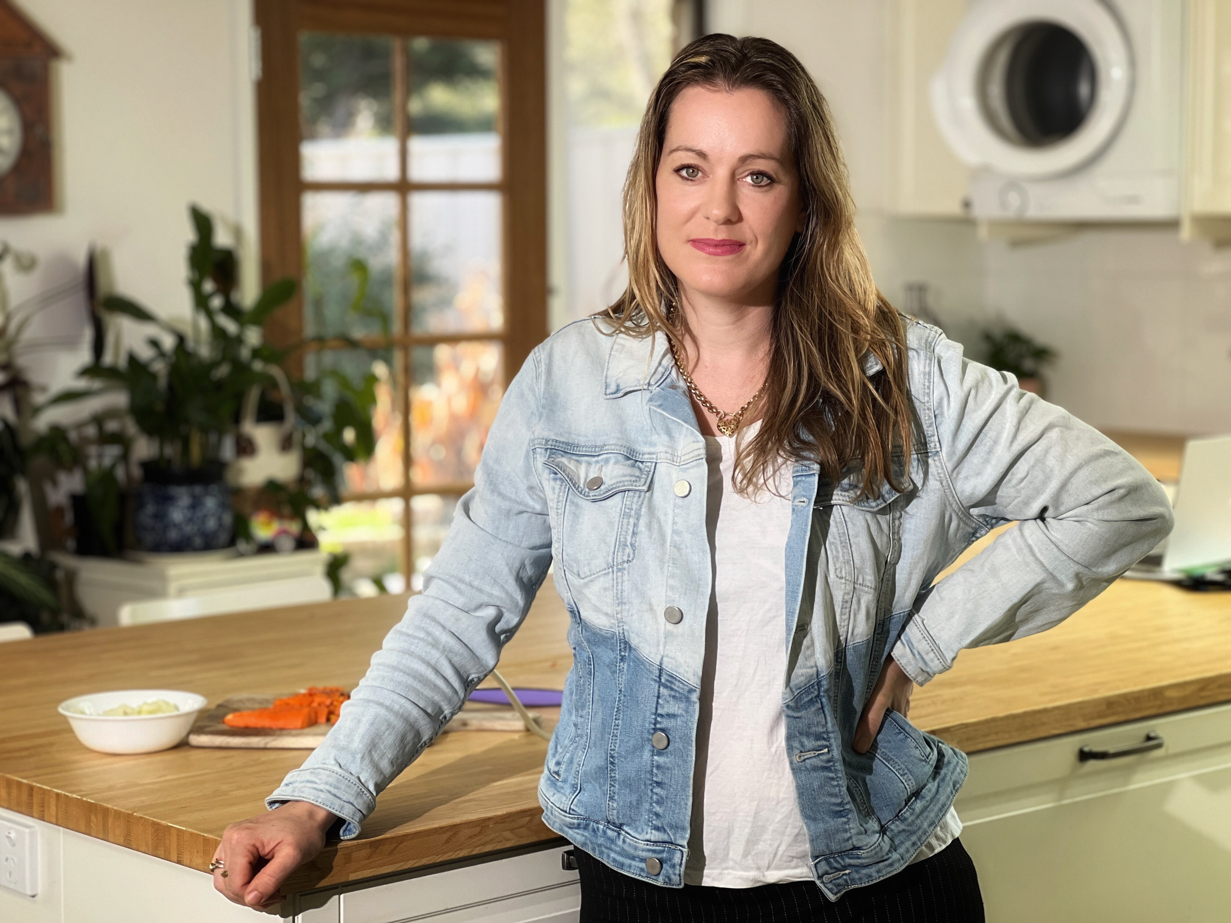 A woman in a two-tone denim jacket, stands at her kitchen bench.