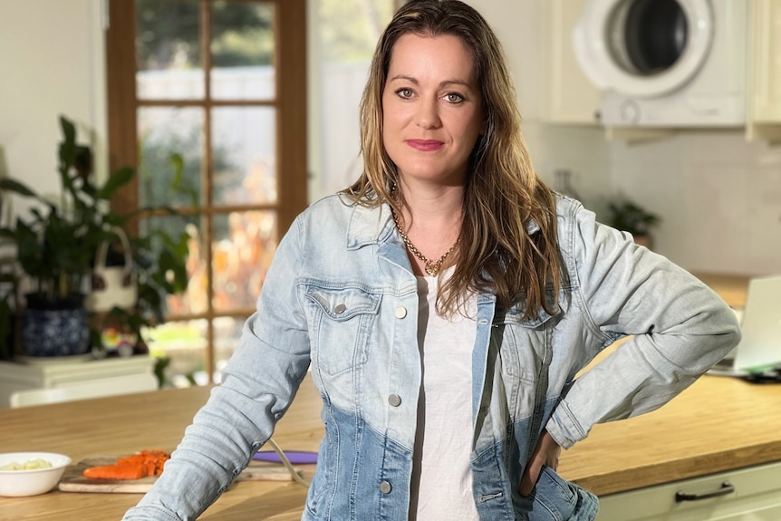 A woman in a two-tone denim jacket, stands at her kitchen bench.