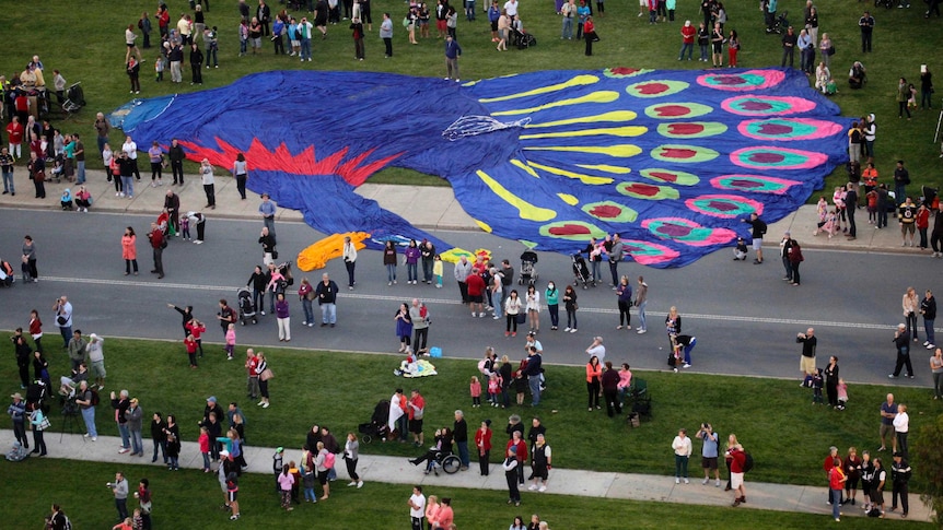 Spectators walk past an uninflated hot air balloon shaped like a peacock.
