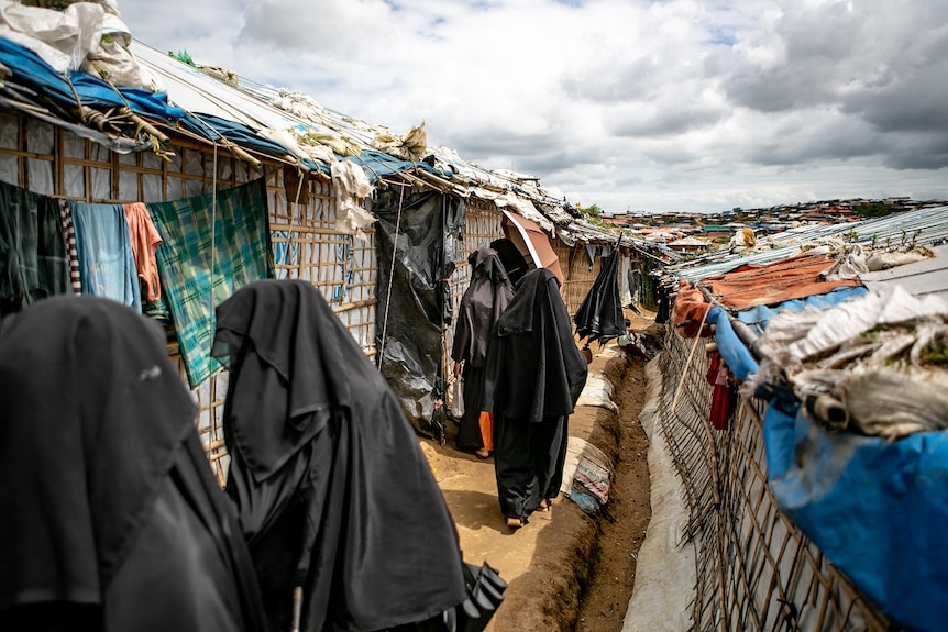 Women in full face and body coverings walk single file through a crowded camp of shacks.