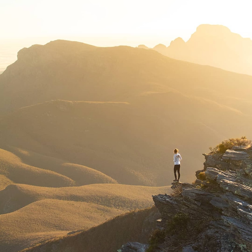 A person stands on a cliff at sunrise.