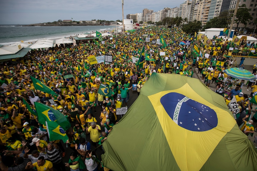 A Brazilian flag covers a section of a large cloud dressed green and yellow gathered between a beach and a row of towers.