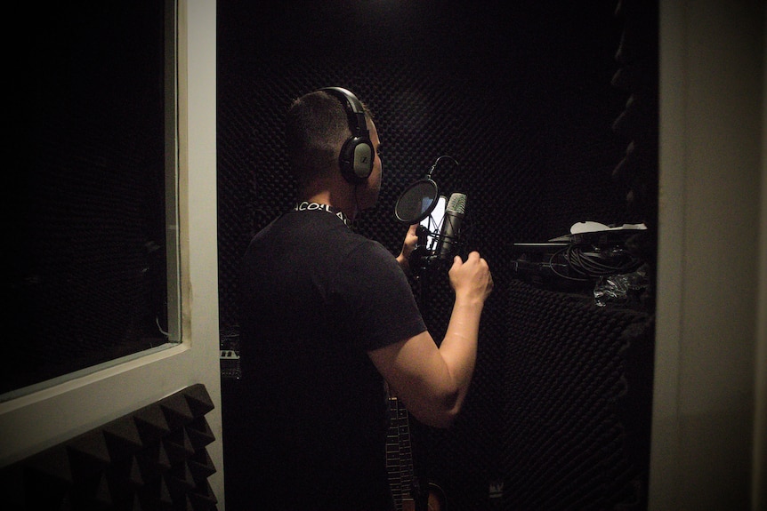 A young man in a black t-shirt wears headphones inside a recording studio.
