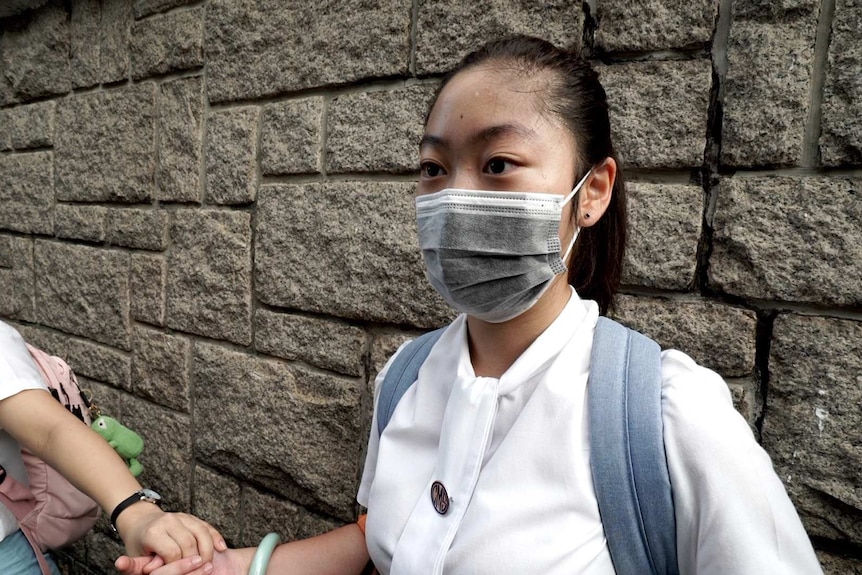 A girl in school uniform, wearing a face mask and backback, holds hands with girls either side of her.
