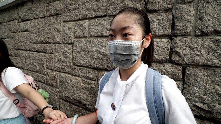 A girl in school uniform, wearing a face mask and backback, holds hands with girls either side of her.