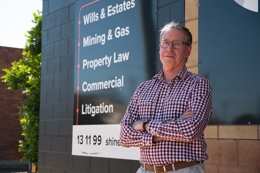 A middle-aged man stands with arms folded outside an office