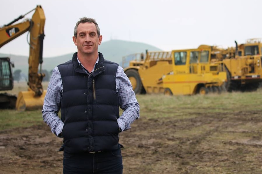 Rohan Arnold stands in a field with some heavy machinery in the distance behind him