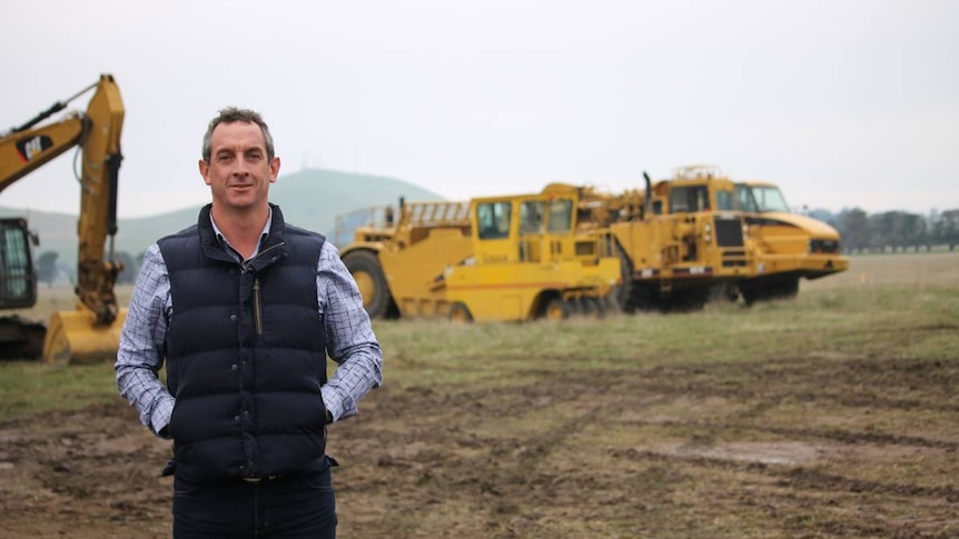 Rohan Arnold stands in a field with some heavy machinery in the distance behind him