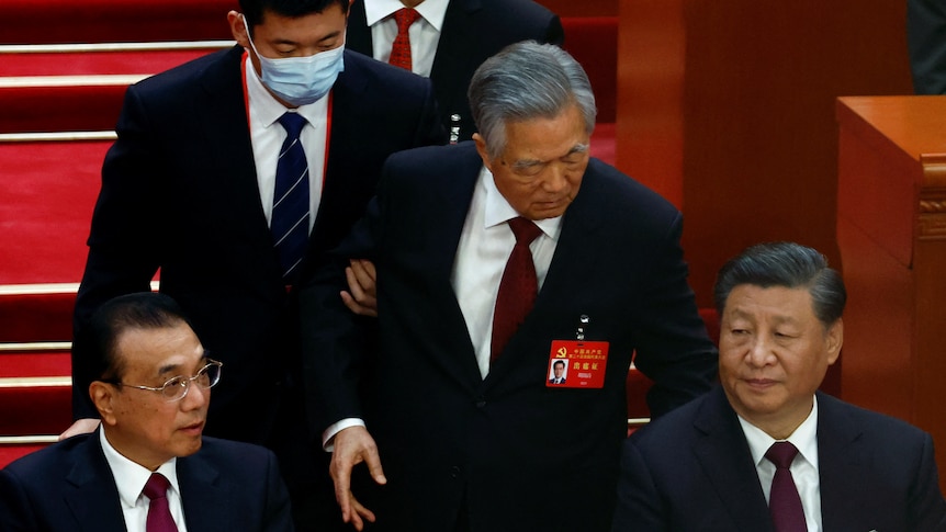 Former Chinese leader Hu Jintao's arm is held by usher as he stands behind seated Xi Jinping.