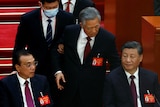 Former Chinese leader Hu Jintao's arm is held by usher as he stands behind seated Xi Jinping.