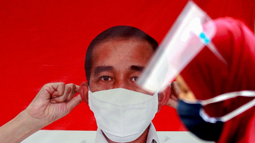 A woman wearing a protective face mask and a face shield walks past a banner depicting Indonesian President Joko Widodo.