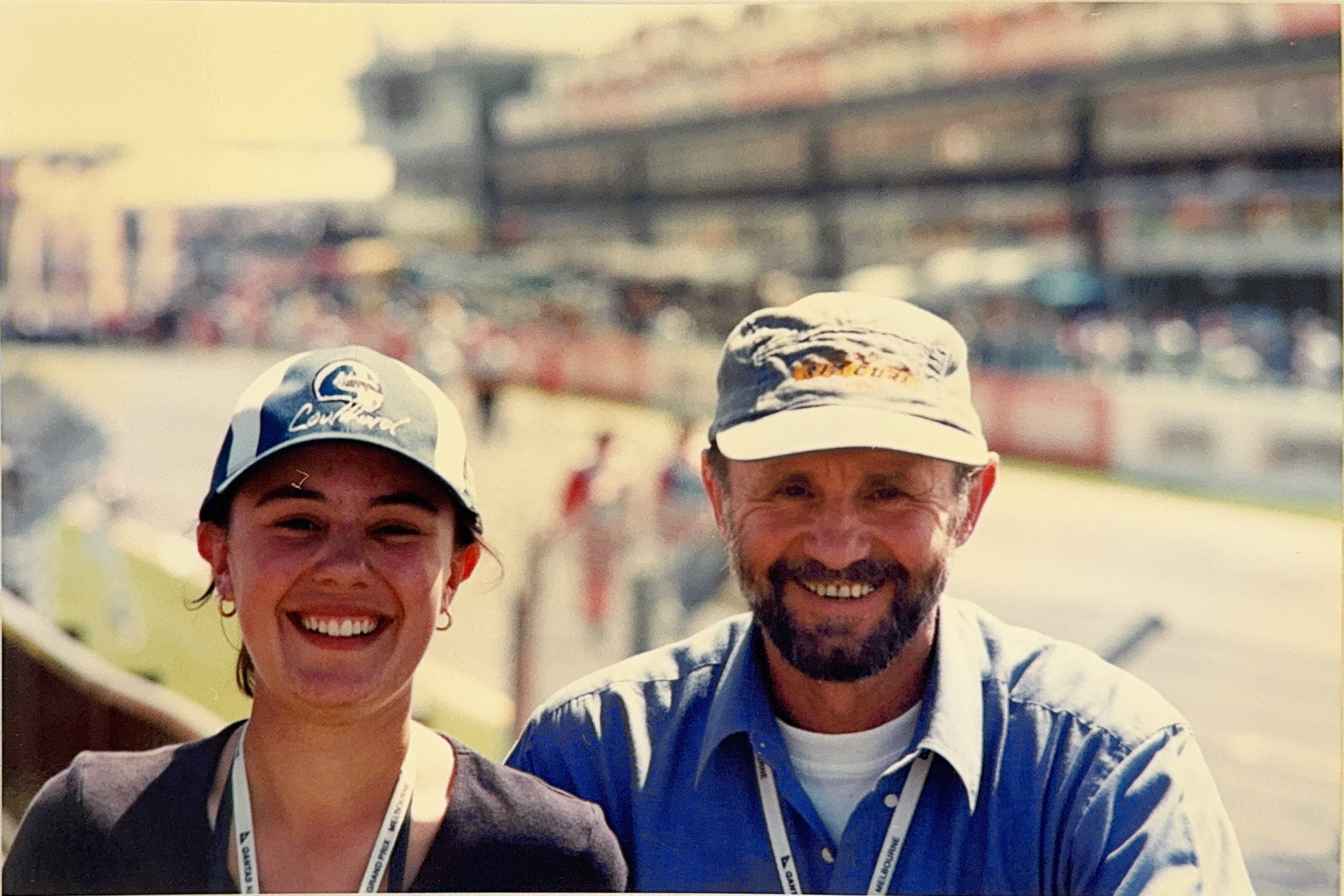 A teenage girl and her father wear caps, smiling big. Behind is out-of-focus car racing track and crowd
