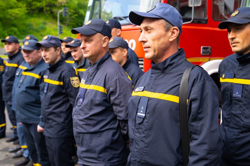A row of emergency workers in navy windbreakers and caps stand in a row in front of a fire truck