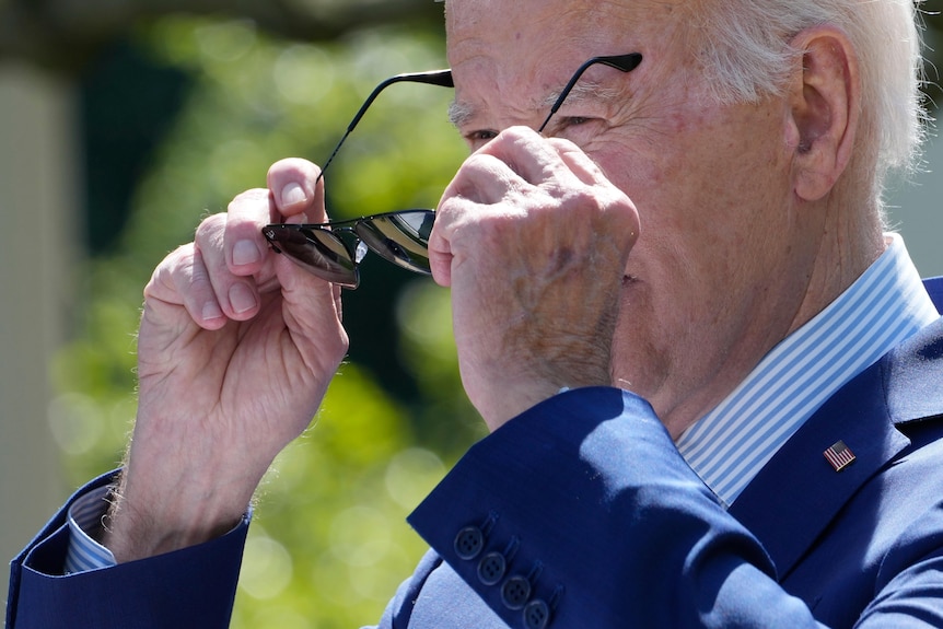 Head and shoulders shot of Joe Biden puting a pair of dark-lensed, black sunglasses up to his face.