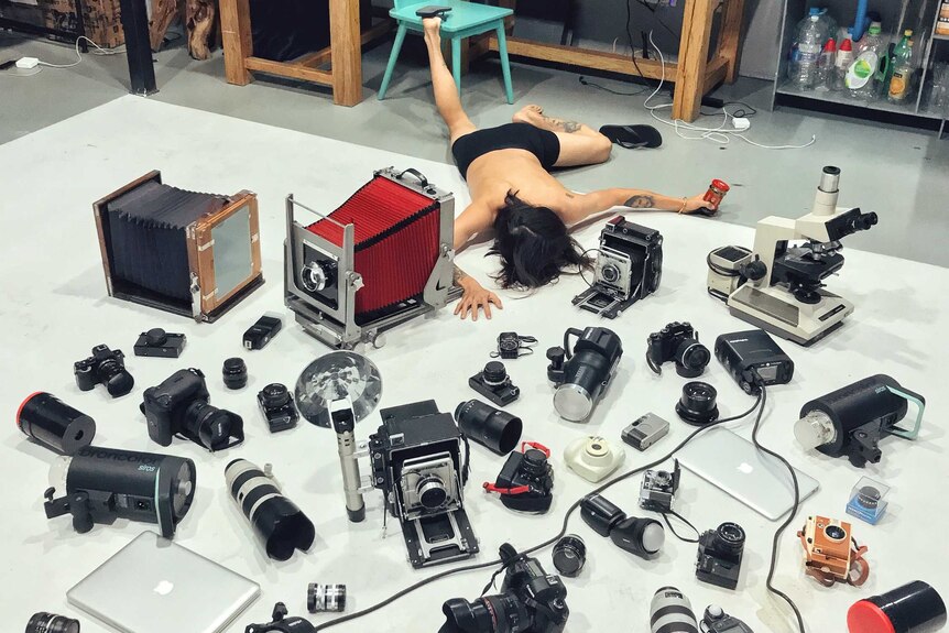 A photographer posed for falling over with many cameras and lenses on the floor of a room where machines and tools at the back.
