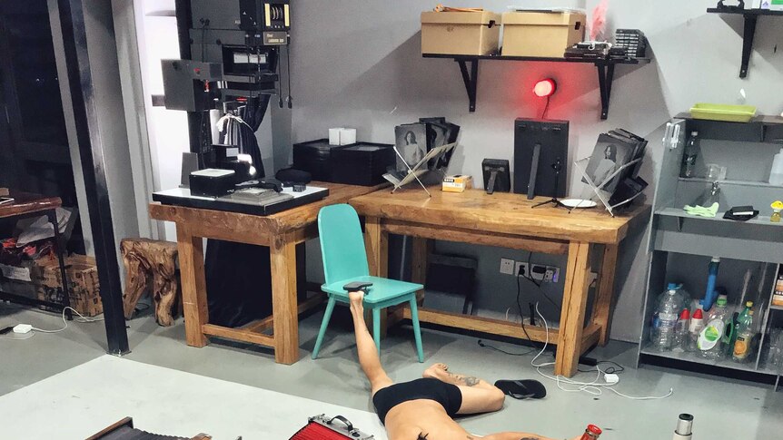 A photographer posed for falling over with many cameras and lenses on the floor of a room where machines and tools at the back.