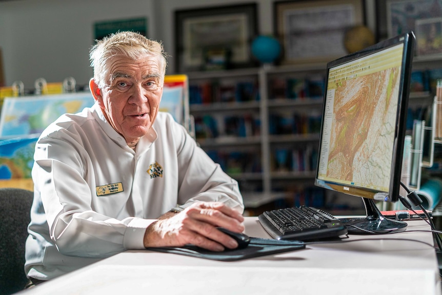 An older man with white hair in a white button-up shirt sits at a computer, maps in the background.