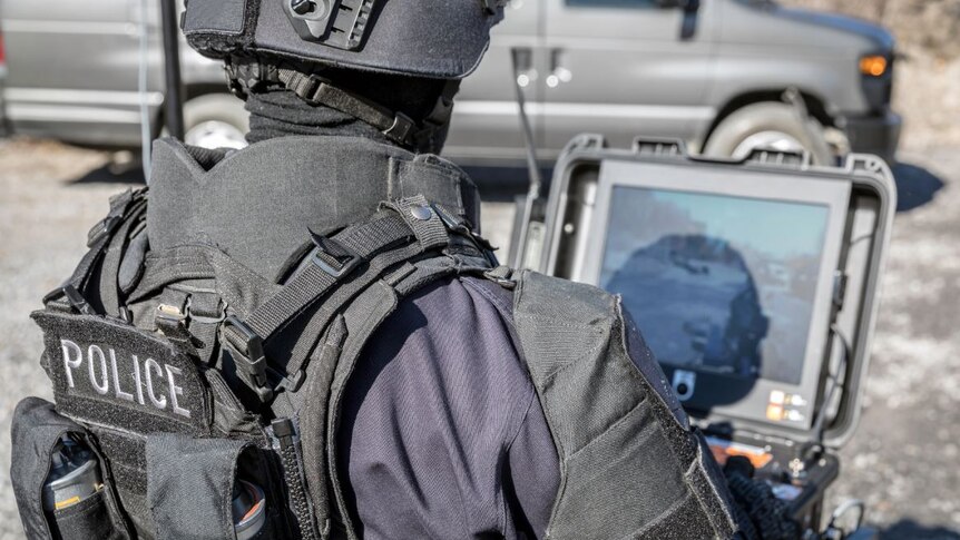 A police officer dressed in helmet and protective padding is outside looking at a computer screen.