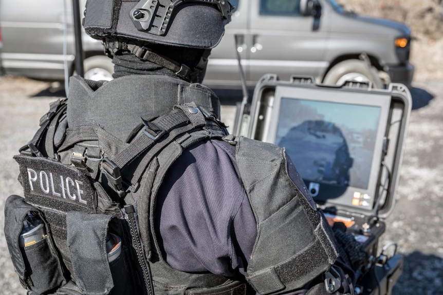 A police officer dressed in helmet and protective padding is outside looking at a computer screen.