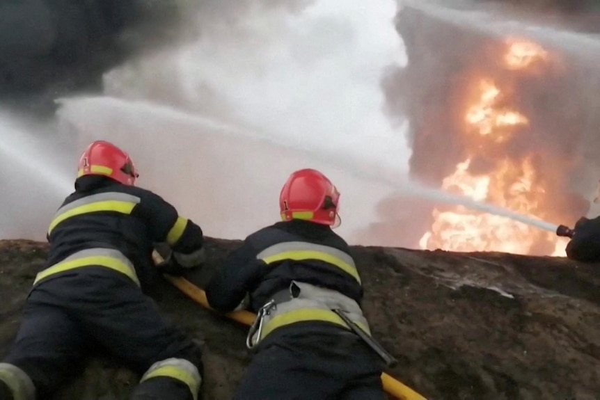Two firemen in uniforms and hard hats lie on a rock, holding a hose which is shooting water into a cloud of smoke and fire.