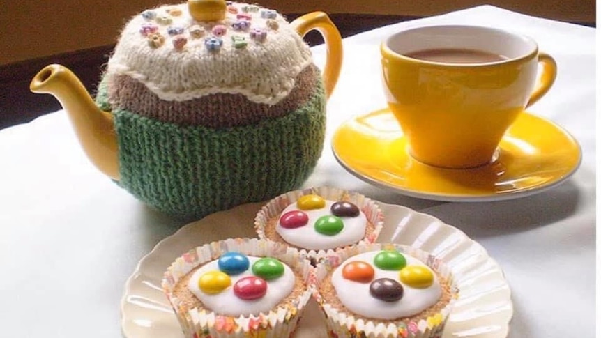 A bright yellow tea cup and tea pot on a table next to three cupcakes with white icing and smarties on them.