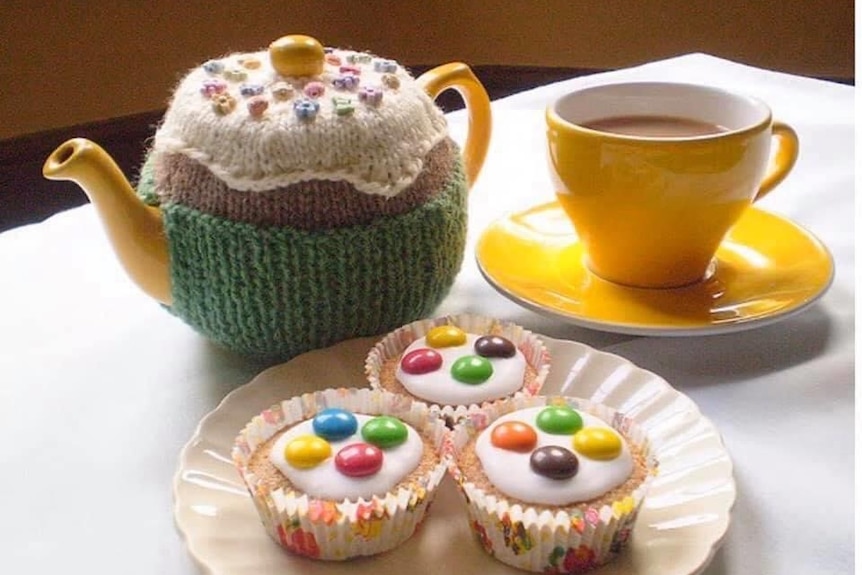 A bright yellow tea pot and tea cup on a table with a plate of three iced cupcakes in front of them.