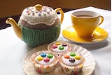 A bright yellow tea cup and tea pot on a table next to three cupcakes with white icing and smarties on them.