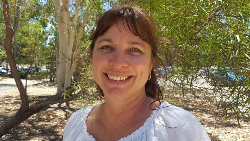 A woman with brown hair, wearing a white shirt, standing in front of some gum trees.