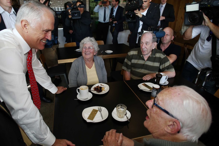 Malcolm Turnbull, surrounded by media, leans on a table in a cafe as he speaks with elderly people seated at the table.