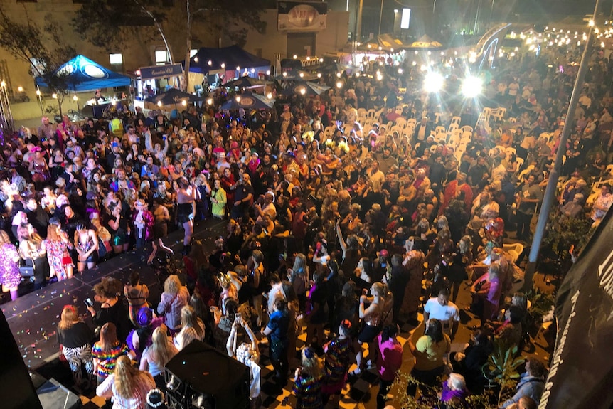 A large and colourful crowd dance in front of a stage.