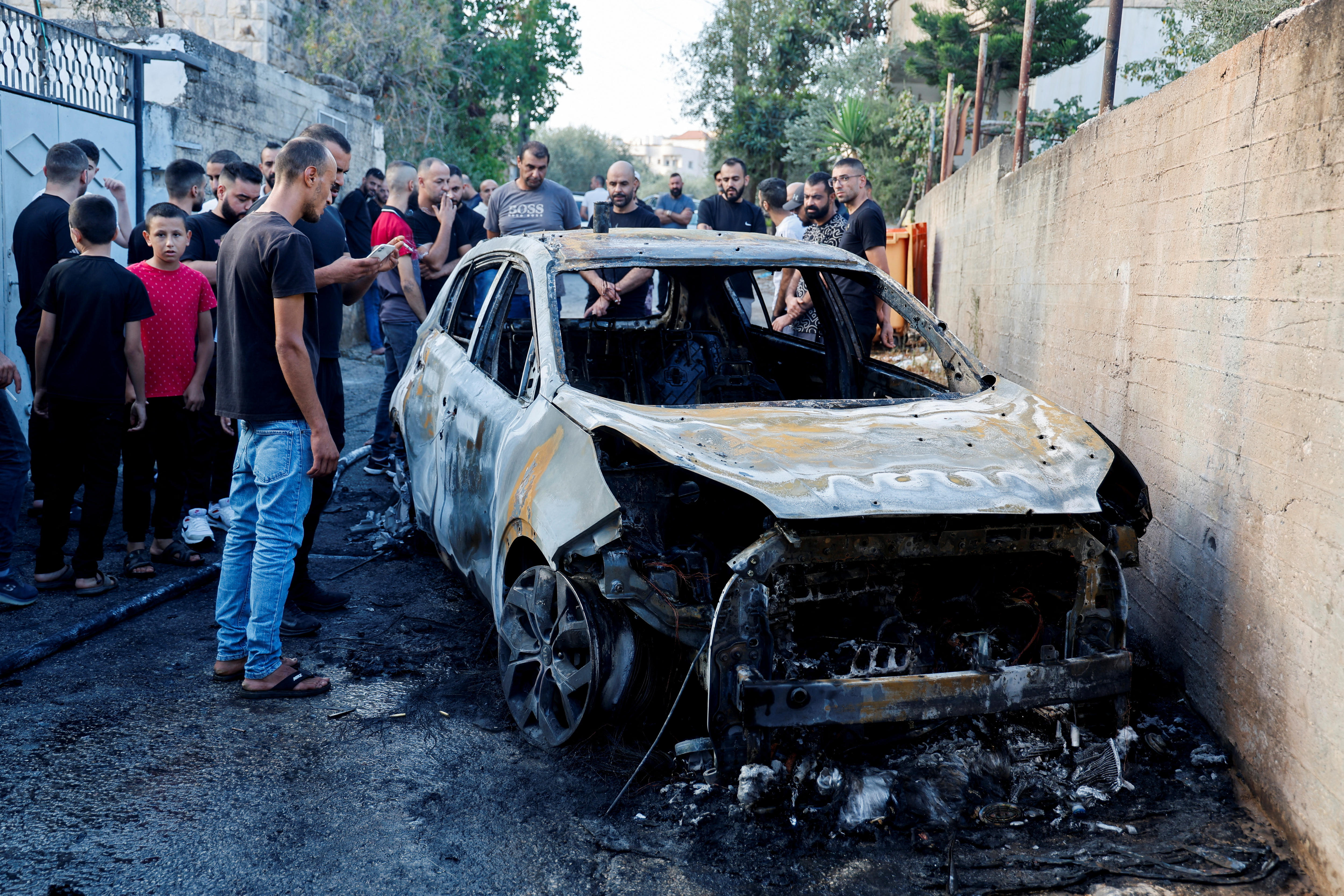 A group of young Palestinian men stand in a street next to the burnt-out wreckage of a car.