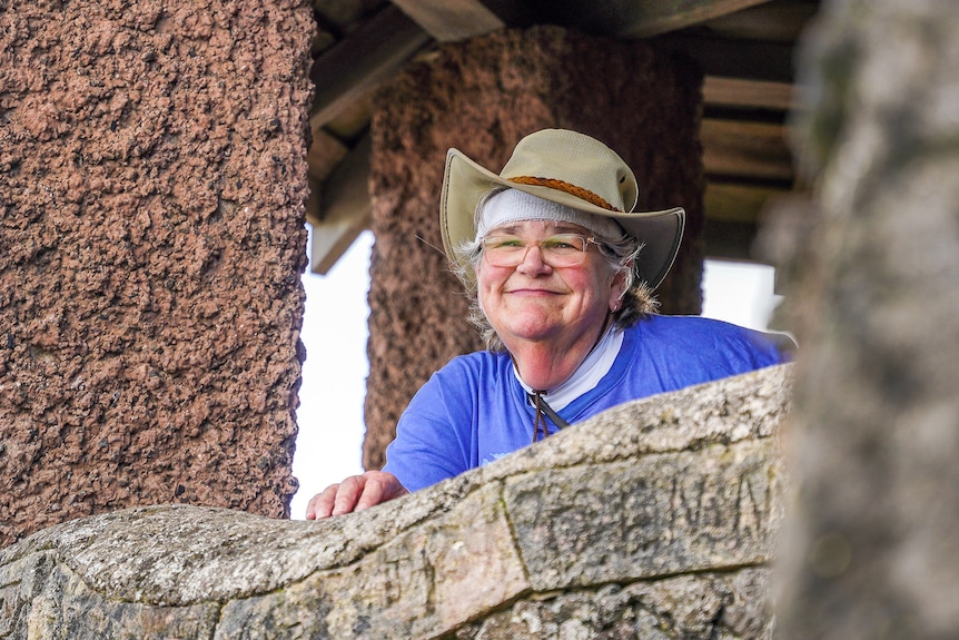 A woman wearing a blue shirt and wide brown hat leans against a concrete hut smiling.