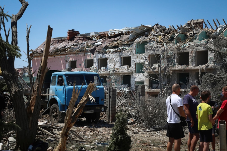 Four people stand outside damaged residential building.