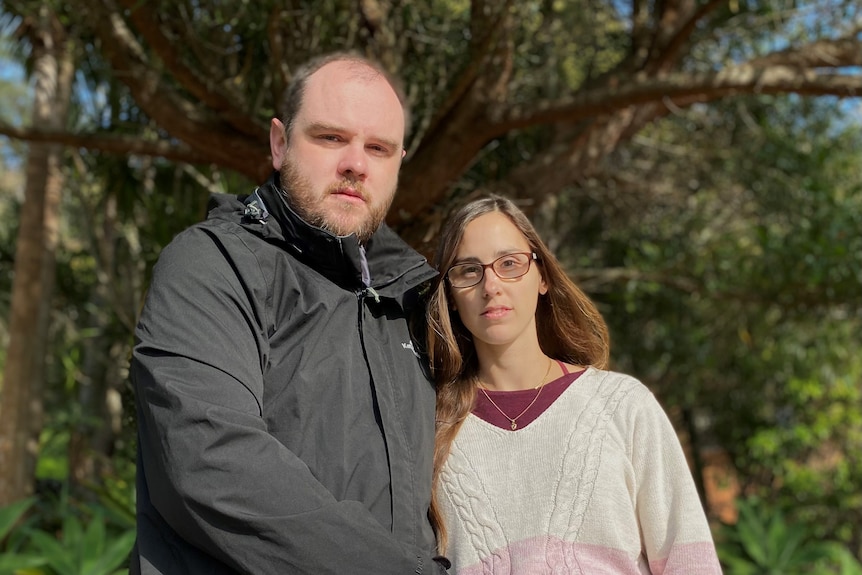 A man and pregnant woman stand under a tree with a sad look on their face