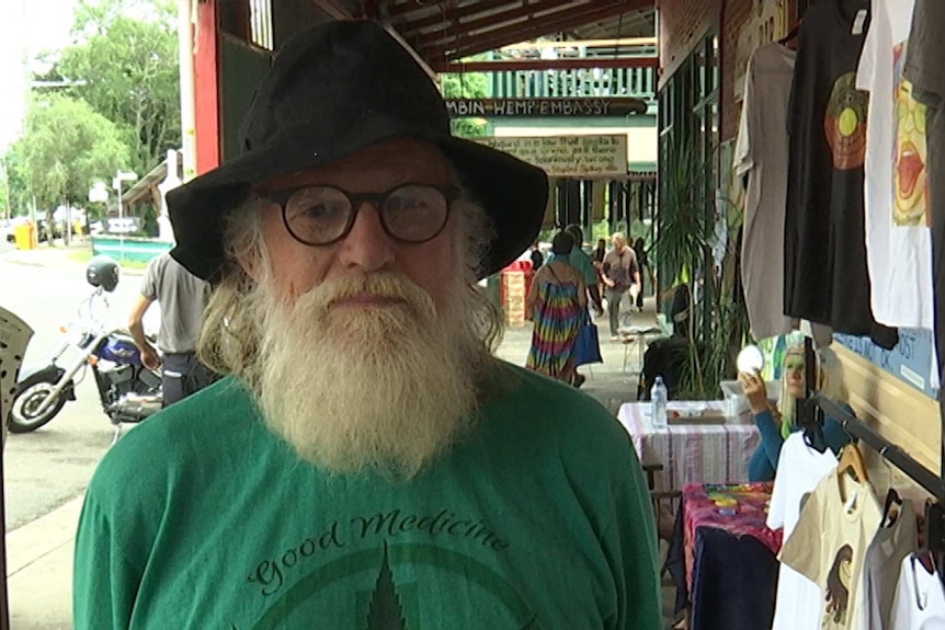 An older man with a white beard and glasses, standing on the street wearing a battered hat and a green t-shirt.