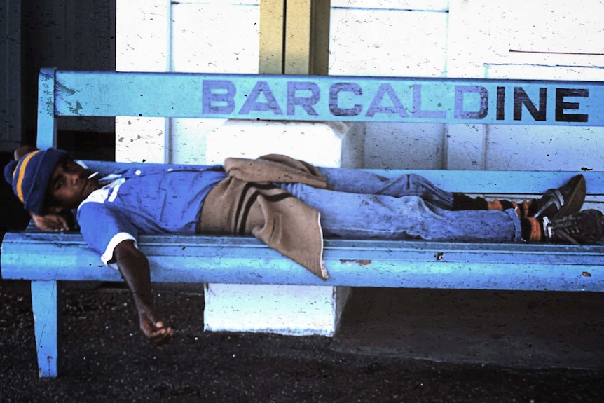 Aboriginal boy lays on a public bench with a cigarette in his mouth.