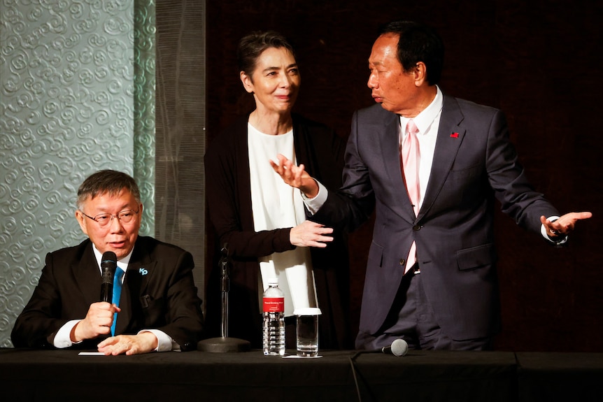 A man sits on stage speaking into a microphone next to a woman and a man standing.