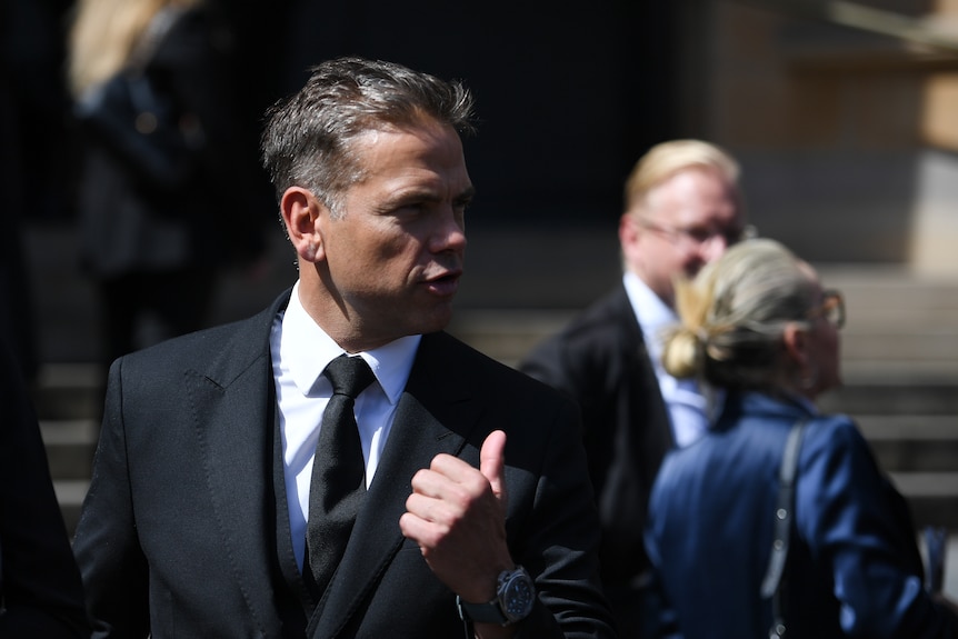 A man in a black suit raises his left hand with his thumb pointing out